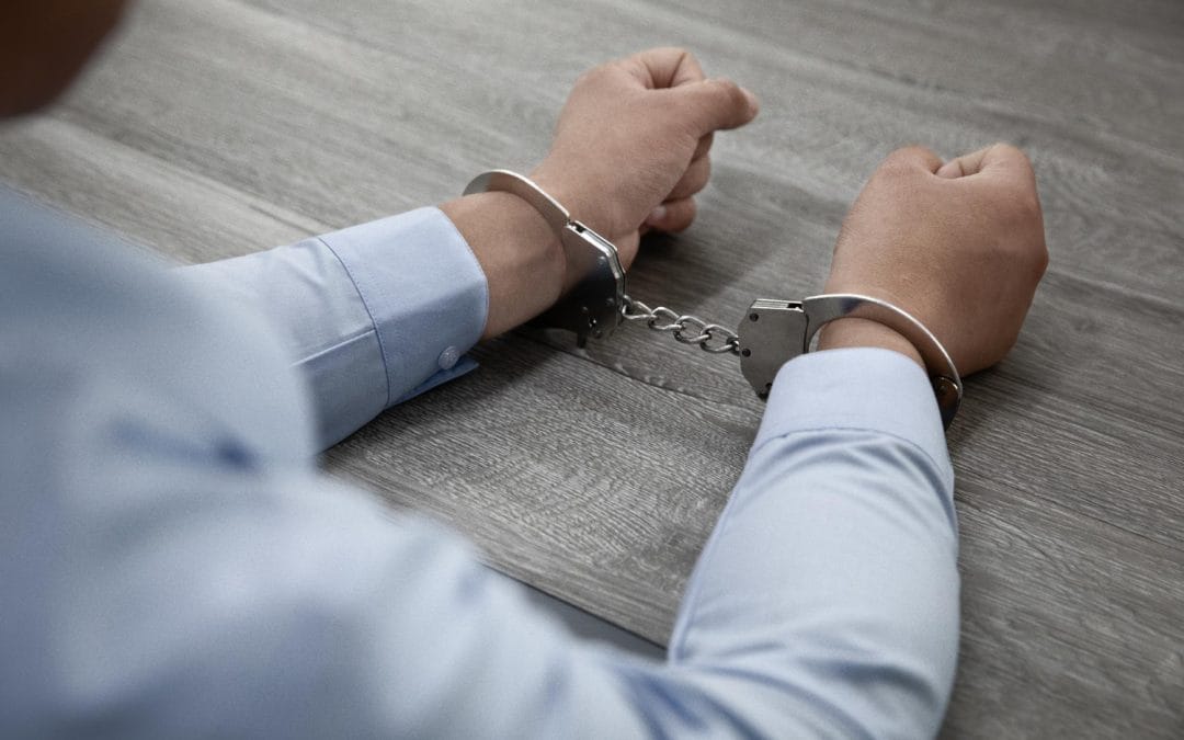 Person in a blue shirt with hands in handcuffs, sitting at a gray wooden table.